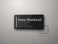 Czarna tabliczka z białym grawerem: pionowa linia, „Panu Markowi”, dedykacja i podpis